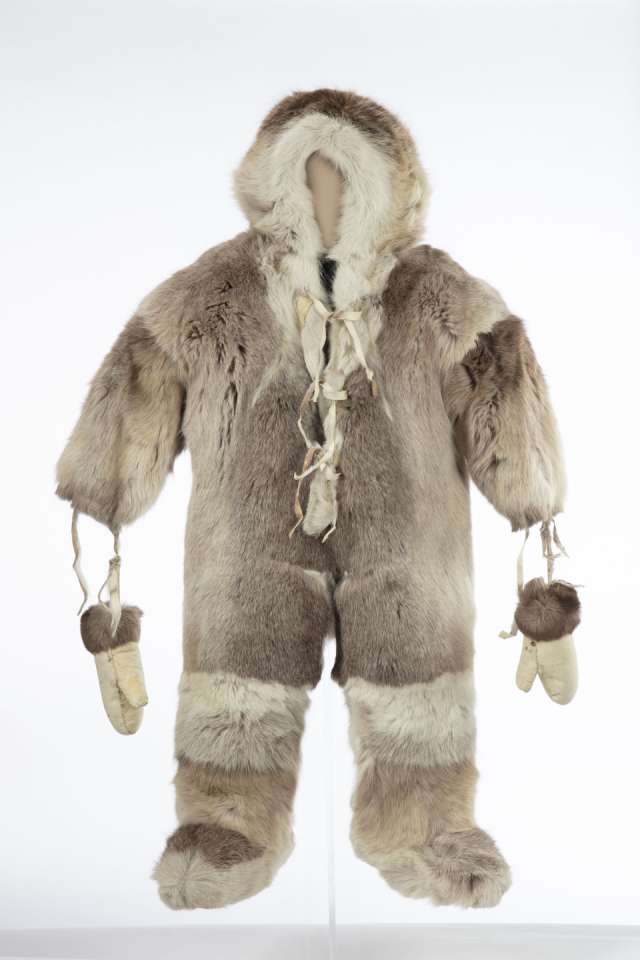 Combinaison en fourrure, vue de face, avec un capuchon. Des mitaines de cuir retourné, ornées de fourrure brune aux poignets, sont suspendues aux manches par des lanières. La fourrure est de couleur plus claire autour du capuchon et au-dessus des bottes qui sont intégrées à la combinaison. La combinaison est fermée par un lacet à l’avant au niveau de la poitrine et du cou.