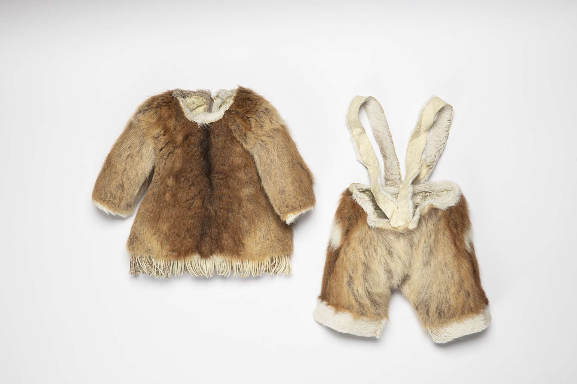 Petit ensemble en fourrure, vu de devant, composé d’un manteau et d’un pantalon à bretelles. Le manteau est doté de franges en bas. Une bande de fourrure blanche borde la taille et le bas de chaque jambe du pantalon, qui est doté de larges bretelles en peau.