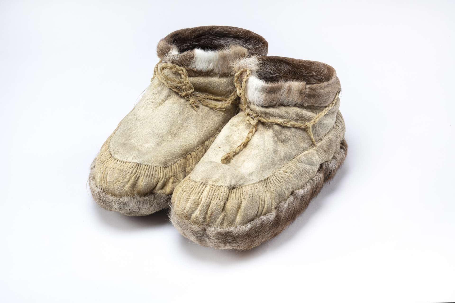 A pair of beige leather ankle boots. It is decorated with brown and white fur around the ankles, has brown fur under the sole, and is equipped with drawstrings.