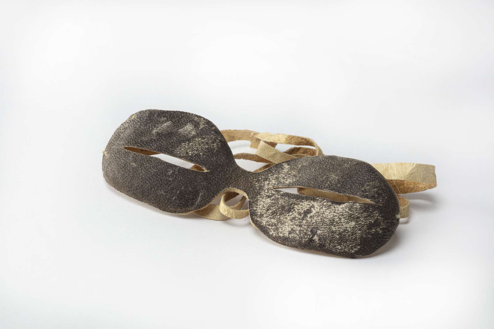 Goggles made of dark brown leather, seen from the front, with two narrow slits for the eyes and a beige leather strap.