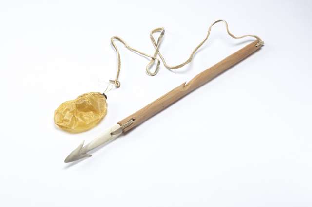 Harpon en bois avec une pointe en matière osseuse blanche, reliée par une lanière de cuir. Une autre lanière de cuir est attachée à un petit flotteur en forme de poche gonflable, jaunâtre.