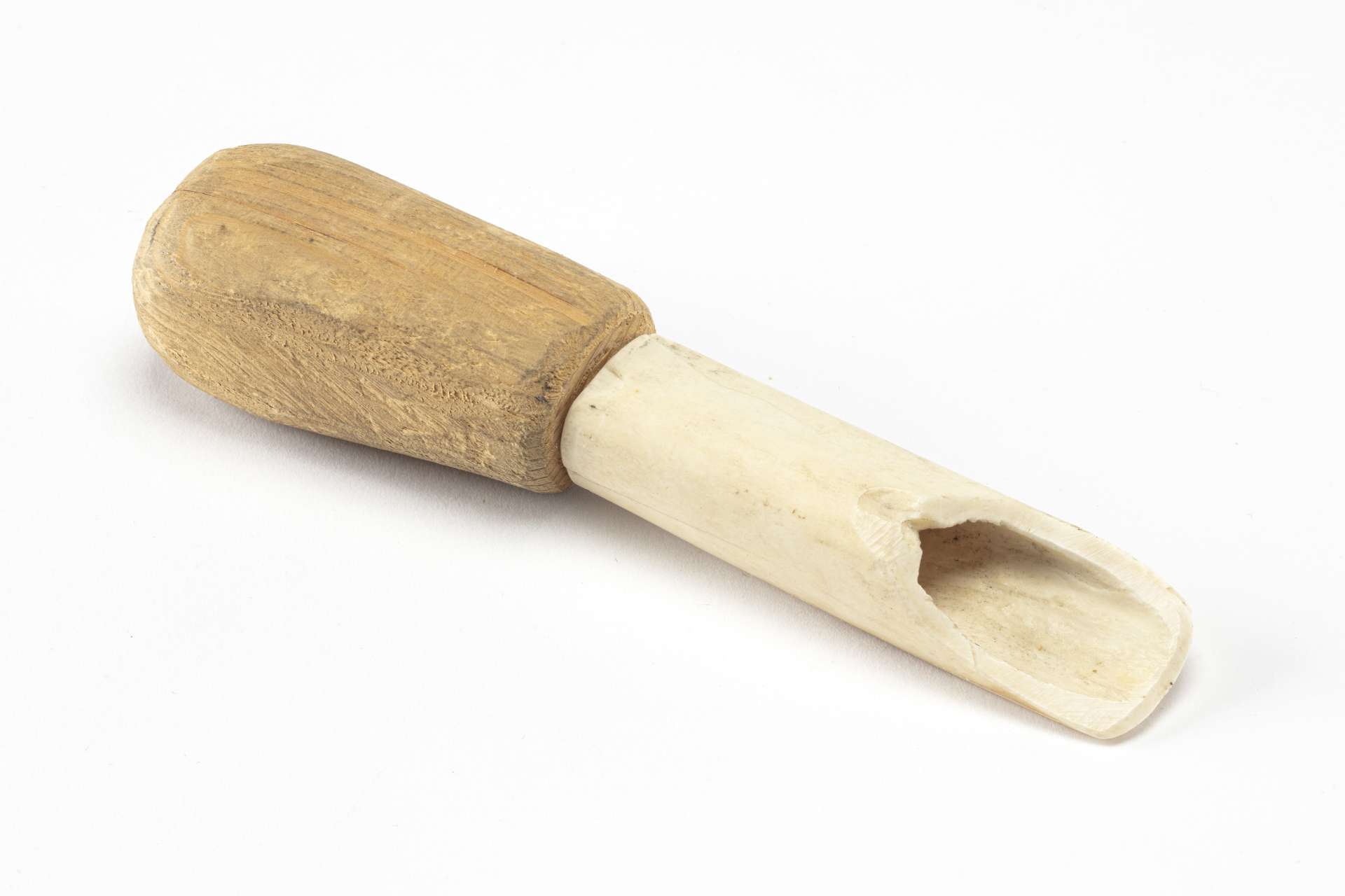 Scraper composed of a handle and a blade. The handle is carved and wooden, rectangular in shape, and slightly rounded on its sides. The blade is hollow and carved from white bone, with a flared lengthwise opening toward its end.