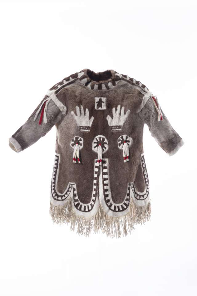 Manteau en fourrure brun ornée de motifs blancs, vu en 360 degrés. Sur la poitrine, deux mains blanches sont représentées sous un petit carré central contenant une silhouette noire. Des motifs circulaires agrémentés de lanière de fourrure et de tissu aux couleurs blanche et rouge sont visibles à l’avant et à l’arrière du manteau. Le dos présente également des motifs linéaires blancs. Le bas du manteau, le col et les épaules présentent des bandes de fourrure brune et blanche alternées. Le bas du manteau est bordé de longues franges.