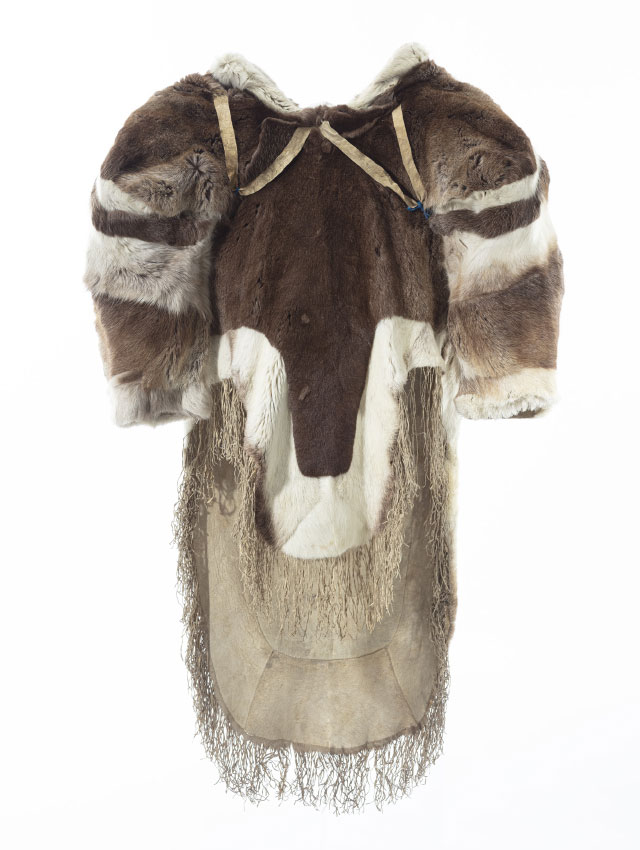Manteau en fourrure vu en 360 degrés, présentant des épaules larges et un grand capuchon. La fourrure brune et blanche est agencée en bandes sur le capuchon et les épaules. Des sangles en forme de « V » sous les épaules sont visibles sur l’avant du manteau. Le pan avant du manteau est plus court que le pan arrière, qui sont tous deux bordés de fourrure blanche et décorés de franges au bas.