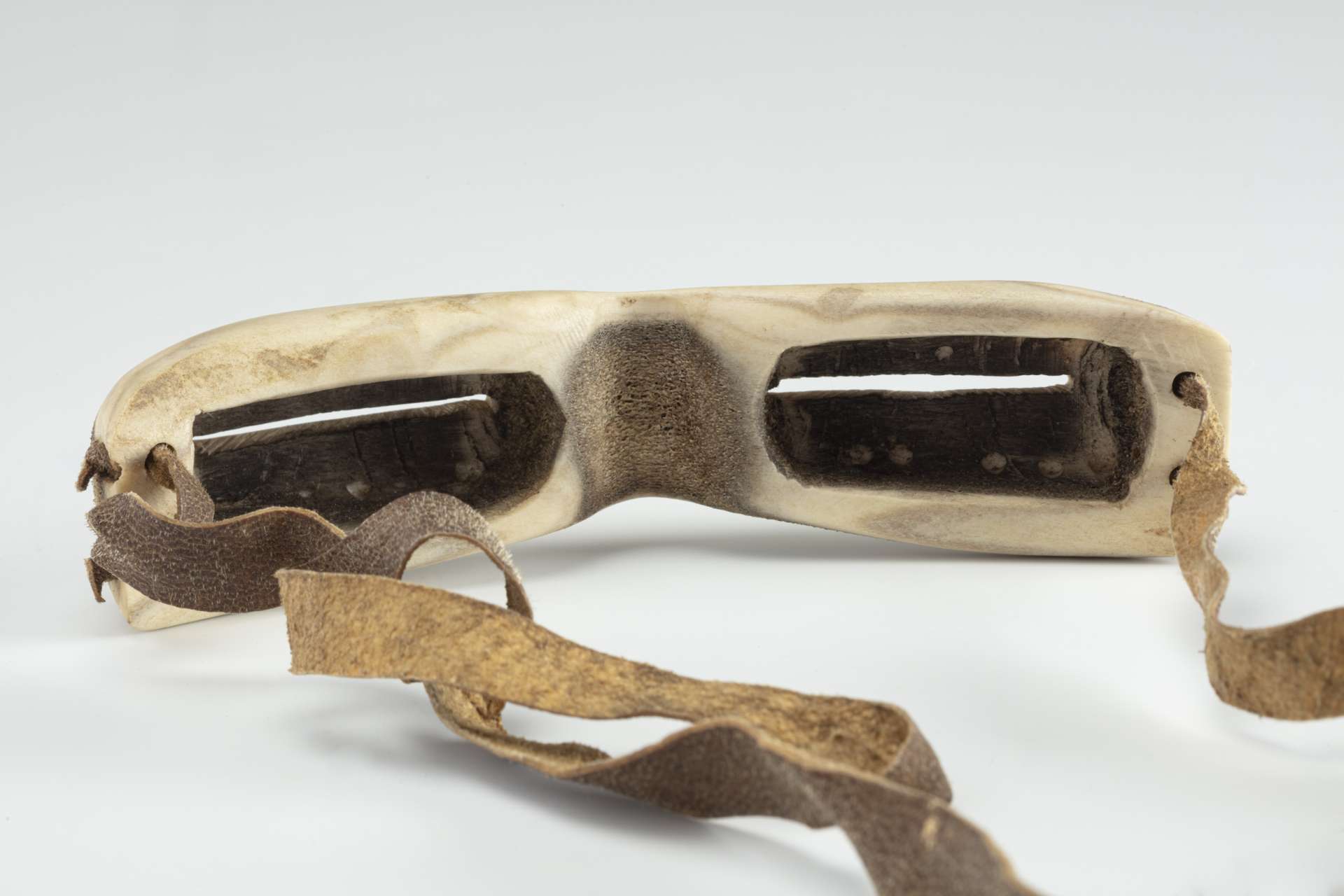 Goggles carved out of white bone matter, seen from behind, showing the interior cavities for the nose and the eyes. The cavities have been darkened, and a brown leather strap is attached to each side.